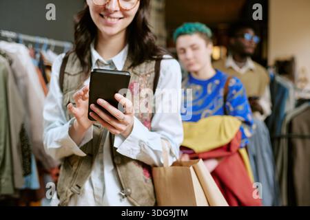 Young Woman Browsing Smartphone While Shopping at Second Hand Store Stock Photo