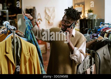 Young Adult Black Man Browsing Second Hand Clothing While Talking on Smartphone Stock Photo