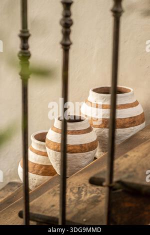 Three handcrafted clay vases with white horizontal stripes arranged on a wooden ledge in a rustic interior setting. Stock Photo