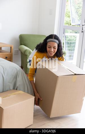 Female teenager kneeling while lifting large cardboard box in bedroom Stock Photo