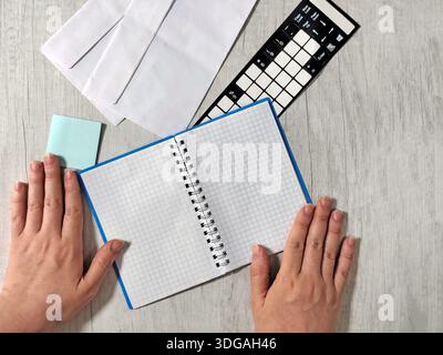 Hands holding an open spiral notebook on a light wooden table with envelopes, sticky notes, and a keyboard sticker sheet. Workspace or study concept. Stock Photo