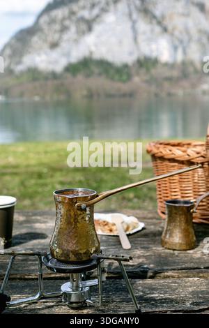 Copper cezve brewing coffee on portable stove outdoors. Picnic setting with wicker basket near lake mountain background. Traditional brewing method du Stock Photo
