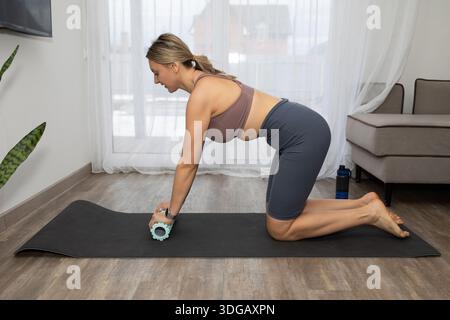 A woman in a sports bra and shorts kneels on a black yoga mat at her home. She is using a green foam roller on her arm. A water bottle is near the mat. Stock Photo