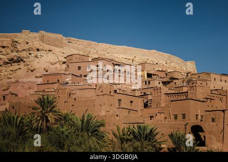Ancient adobe fortress village in arid desert landscape with palm trees, traditional earthen architecture and mountain backdrop under clear blue sky. Stock Photo