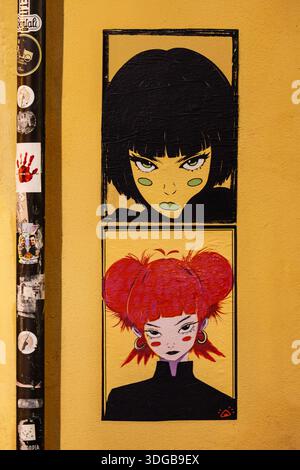 Colorful urban street art featuring two stylized female anime characters posted on a textured yellow wall next to a decorated pipe Stock Photo