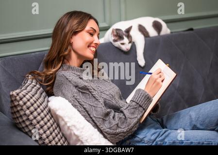 Cheerful cozy dressed woman sitting on sofa and writing diary. Her cat is lying beside her. Stock Photo