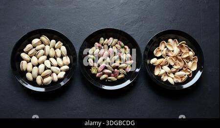 Three black bowls of pistachios, one with shelled pistachios, one with shelled pistachios, and one with empty shells, isolated on a black background. Stock Photo