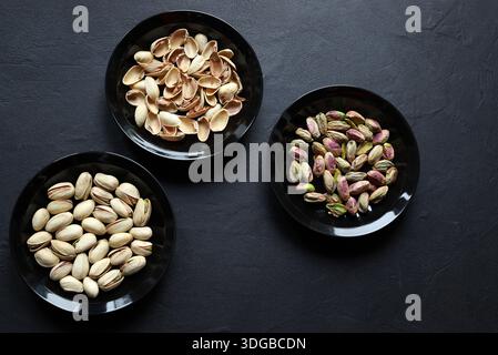 Three black bowls of pistachios, one with shelled pistachios, one with shelled pistachios, and one with empty shells, isolated on a black background. Stock Photo
