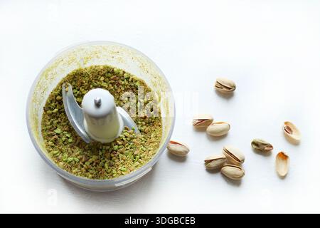 Pistachio kernels in a food processor and some whole, unshelled pistachios next to them on a white background. Overhead view. Stock Photo