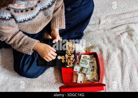 Top view of a young woman sitting on a bed while counting euro coins in an open red cash box filled with euro banknotes Stock Photo