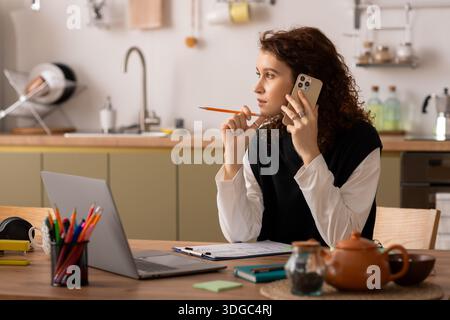Curly-haired woman chats on the phone, deep in thought, surrounded by a warm indoor environment. Stock Photo