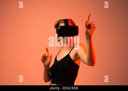 Female user wearing VR headset and interacting with virtual interface using hand gestures in futuristic red neon studio lighting Stock Photo