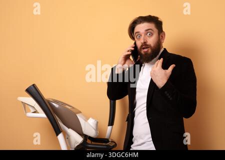 Surprised businessman in suit talking on smartphone next to treadmill against orange background, blending work and fitness. Stock Photo