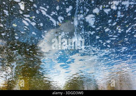 A lake background with patches of foam reflecting a blue sky, with clouds and autumn trees distorted by gentle ripples. An inverted water mirror, an a Stock Photo
