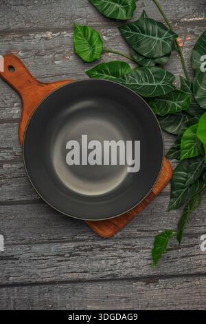 A black bowl sits on a wooden cutting board with green leaves on top. The bowl is empty and the cutting board is made of wood. Concept image Stock Photo