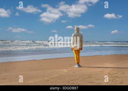 A solitary woman figure stands on a wide sandy beach, gazing out at rolling ocean waves beneath a bright blue sky dotted with soft clouds Stock Photo