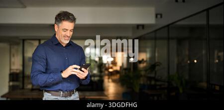 Smiling mature business man corporate leader using cellphone standing in office. Stock Photo