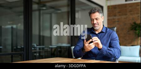 Smiling mature business man corporate leader using cellphone sitting in office. Stock Photo