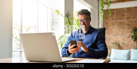 Happy middle aged businessman using mobile phone working in office. Stock Photo