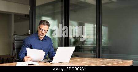 Busy mature professional business man using laptop reading document at work. Stock Photo