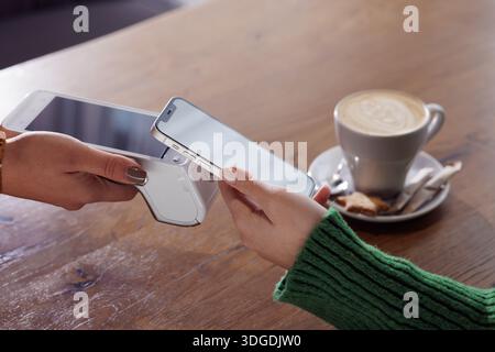 Woman paying with smartphone using contactless terminal in cafe, latte and dessert on wooden table. Stock Photo