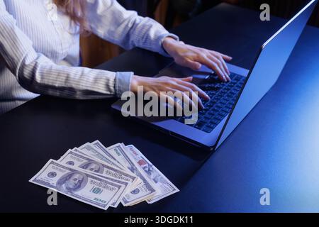 A person typing on a laptop with a stack of cash on the table. The scene is dimly lit, highlighting the hands and the money, suggesting a focus on fin Stock Photo