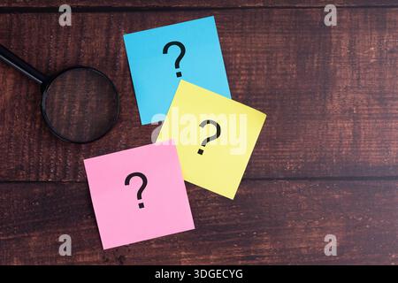 Colorful sticky notes with question marks and magnifying glass on wooden desk. Stock Photo