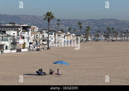 People relax under a beach umbrella on the sand at Newport Beach, California, with oceanfront homes and palm trees lining the shore. Stock Photo