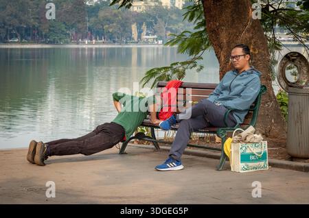 Life on a park bench. One Vietnamese man exercises while another sits impassively next to Hoan Kiem lake in central Hanoi, Vietnam. The men are unknow Stock Photo