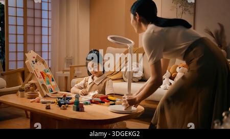 Asian mother placing LED desk lamp for daughter studying at wooden table in evening Stock Photo