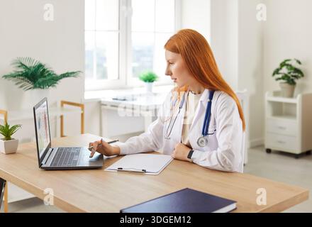 Female doctor sitting in front of laptop, writing report or prescription after patient examination. Stock Photo