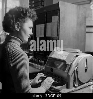 Slaviansk, Ukrainian SSR, Soviet Union - Spring 1960s: A black and white archival medium shot of a female employee operating a teletype or dispatch console at a communication center. The woman, wearing a ribbed turtleneck sweater and short curly hair, is typing on a keyboard with a focused expression. Behind her, a complex switchboard panel is visible. This authentic vintage photograph captures the atmosphere of Soviet labor, women in the workforce, early technology, and the peaceful daily life in the industrial city of Donbas during the mid-20th century. Stock Photo