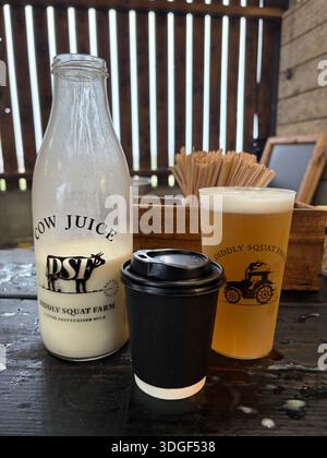 Fresh farm milk bottle, takeaway coffee cup and craft beer on rustic wooden table at countryside Diddly Squat farm café Stock Photo