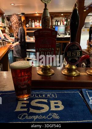 Pint of beer on a pub counter with traditional British ale pump handles and bartender in the background inside a classic English pub. Stock Photo