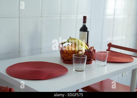 bright minimalist kitchen table set for two with red placemats, fruit bowl centerpiece and wine bottle, cozy home dining and contemporary lifestyle co Stock Photo