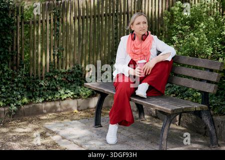 Mature woman in red trousers and white jacket sitting on a wooden park bench. She has red headphones and holds a pink reusable coffee cup. Stock Photo