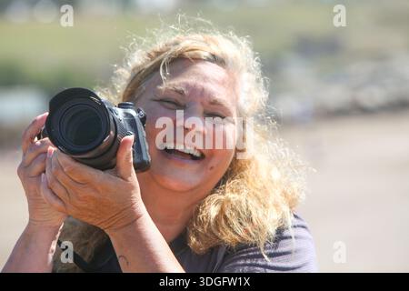 A smiling mature woman holding a camera, taking photos at a busy beach on a bright summer day. The image embodies active aging, leisure, travel, and a Stock Photo