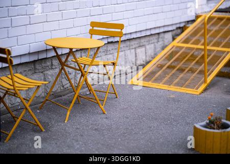 Two yellow metal chairs and a matching round table stand on asphalt against a white brick wall, next to a yellow metal ramp or staircase, with a yello Stock Photo