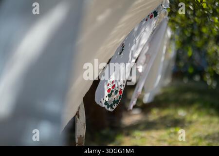 White tablecloths, some embroidered, hang to dry on a long clothesline with clothespins in a fruit orchard, with leaf shadows and green foliage in the Stock Photo