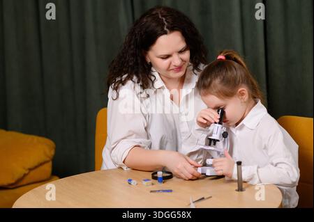 Mom and daughter examine specimens under microscope while sitting in living room. Woman helps girl conduct hands-on scientific experiments using micro Stock Photo