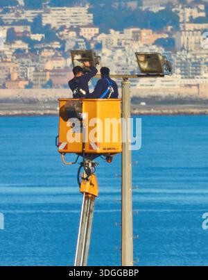 Two Workers In A Yellow Bucket Lift Inspecting A Light Pole Over The Sea With City Background Stock Photo