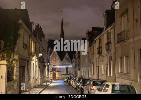 Night view of a narrow street with parked cars and residential buildings leading to a historic stone church tower with festive lights under a dark clo Stock Photo
