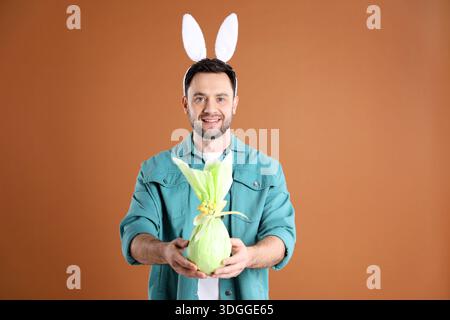Easter celebration. Smiling man in headband with bunny ears holding wrapped gift on brown background Stock Photo