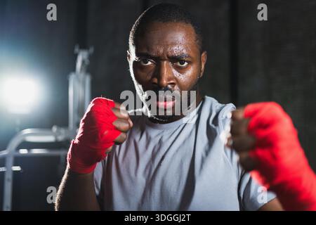 Boxer in gym. Aggressive African man fighter resting after training boxing ready for fight looking at camera. Strong sweated man training punches looking concentrated straight preparing for sparring || Model released Stock Photo