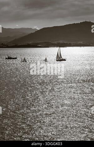 Santander, Spain – Nov 19, 2021: A black and white photograph of a lone sailboat on the glittering water of the Bay of Santander, with hazy mountains Stock Photo