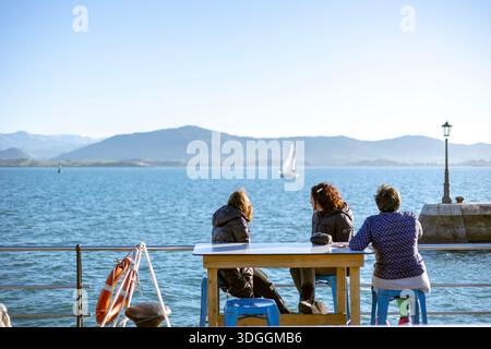 Santander, Spain – Nov 19, 2021: Three women sit at a table on the waterfront promenade in Santander, looking out at the bay with sailboats and mounta Stock Photo