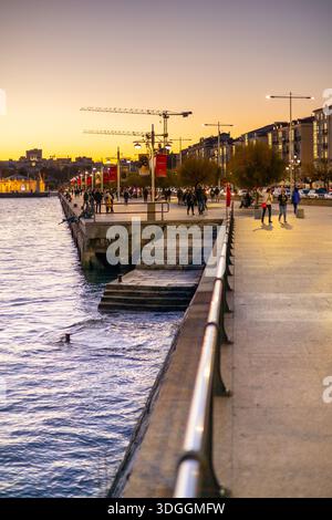 Santander, Spain – Nov 19, 2021: Crowds of people walk along the Paseo de Pereda waterfront promenade at dusk, with the city skyline of Santander illu Stock Photo
