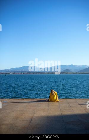 Santander, Spain – Nov 19, 2021: A woman in a yellow jacket sits alone on the waterfront promenade in Santander, looking out at the bay and distant mo Stock Photo