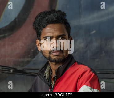A young man in a red and black jacket, captured in a moment of quiet focus against a dark urban wall. His sideways gaze and the subdued background cre Stock Photo
