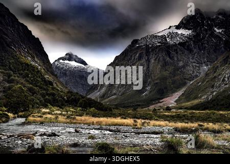Dramatic landscape with snow-capped mountains and a river in the foreground under a cloudy sky, Milford Road, New Zealand Stock Photo
